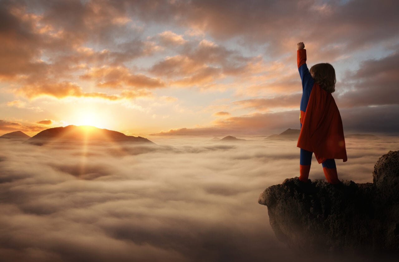 Little boy acting like a superhero on top of the mountain at sunset with copy space